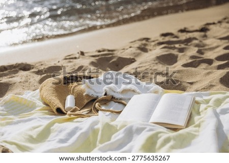 Beach trip essentials including an open book, straw bag with sunglasses and sunscreen, and a white linen hat, lying on a towel near the sparkling ocean waves