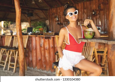 Beach Vacation Concept. Young And Sexy Woman Sitting In Authentic Beach Bar With A Coconut Drink 