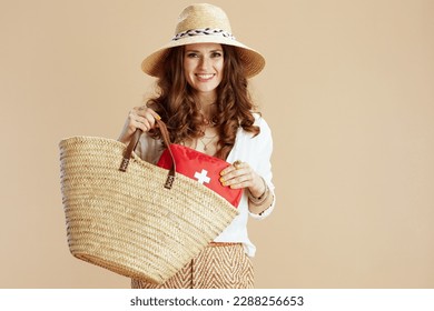 Beach vacation. smiling modern 40 years old housewife in white blouse and shorts against beige background with first aid kit, straw bag and summer hat. - Shutterstock ID 2288256653