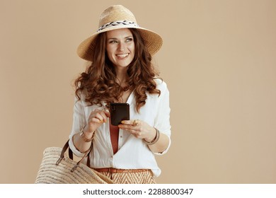 Beach vacation. smiling stylish 40 years old housewife in white blouse and shorts on beige background with straw bag, smartphone and summer hat. - Powered by Shutterstock