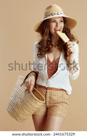 Beach vacation. smiling stylish middle aged woman in white blouse and shorts isolated on beige with straw bag, ice cream and straw hat.