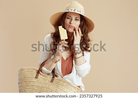 Beach vacation. smiling trendy middle aged housewife in white blouse and shorts on beige background with straw bag, ice cream and straw hat.