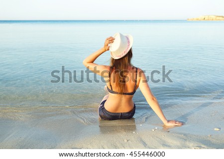 Beach vacation. Young woman in a hat and bikini sitting on the beach - view from the back