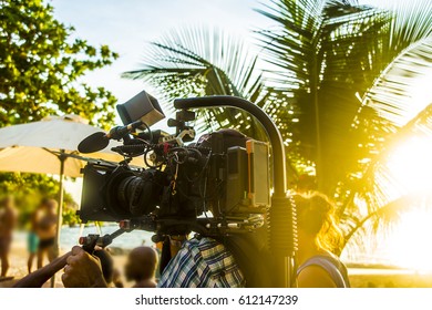 Beach Video Making, Professional Videographer Taking Video Of Young People At Summer Beach Party. Sunny Tropical Day. Sun Rays Beam 