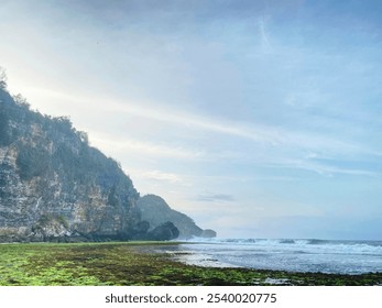 Beach view with rocky cliffs, ocean waves, and green algae-covered shore at Ngrumput beach, Yogyakarta, Indonesia - Powered by Shutterstock