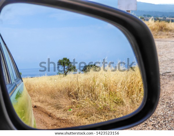 A beach view seen in the side\
mirror of a car. Driving through a graveled road. Ground is\
overgrown with a dried grass. Some trees growing next to the\
beach.