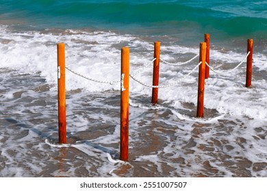 Beach where several wooden posts are embedded in the sand, connected by white ropes. Posts and ropes form a barrier or boundary, with waves washing up against them and the ocean visible in background - Powered by Shutterstock