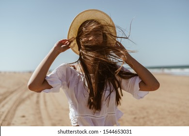 Beach. The wind covered her face with her hair. She holds the hat with her hands.