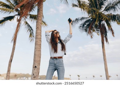 Beach, woman, jeans, white shirt, cell phone a woman in jeans and a white shirt stands on the beach with her arms raised, holding a cell phone, exuding a sense of freedom and joy in a coastal setting - Powered by Shutterstock