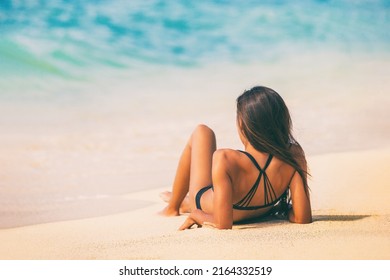 Beach woman lying down on sand relaxing sunbathing on Caribbean travel summer vacation lifestyle. View from behind girl tanning under the sun in black bikini