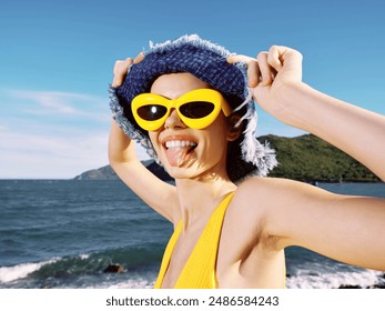 Beach, woman, sunglasses, hat, ocean a woman in sunglasses and a hat on a sandy beach sticks out her tongue playfully with the ocean in the background - Powered by Shutterstock