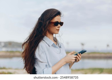 Beach, woman, sunglasses, phone a woman in sunglasses stands on the beach near a body of water, intently looking at her phone while enjoying the serene coastal scenery - Powered by Shutterstock