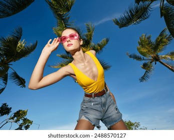 Beach woman in sunglasses and yellow top stands on sandy shore with palm trees and clear blue sky in background, capturing a sunny tropical vacation vibe - Powered by Shutterstock