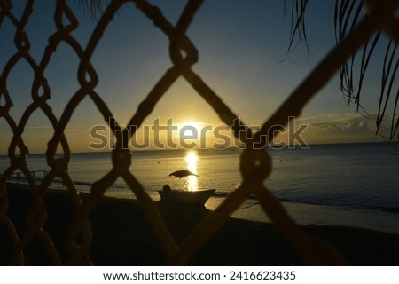 Beachfront silhouette of boat and palm branches at sunset through metal mesh