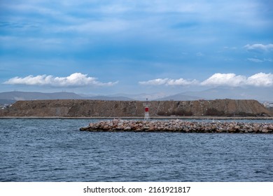 Beacon On Breakwater Rocky Wall Construction At Harbor Entrance Of Greek Island Background. Lighthouse On Seawall, Protection At Ripple Sea, Blue Sky.