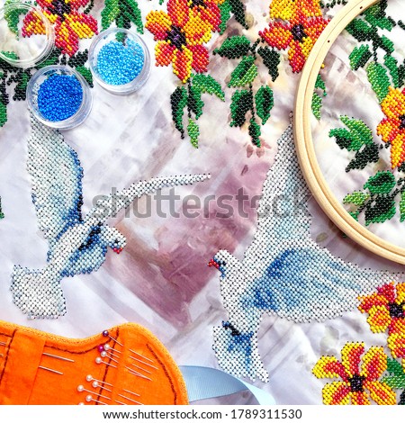 Beads embroidery of dove birds & orange flowers with leaves. Embroidery beads work on table - beads, hoop, needles. Close-up woman embroidery beads work top view two pigeon and flowers pattern