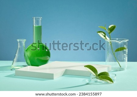 Beaker, petri dish containing green tea leaves, white podium, erlenmeyer flask and flat - bottom florence flask containing green liquid on a blue background. Space for cosmetic advertising.