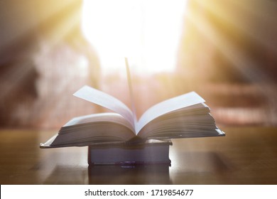 The beam shines down to the open magic book on wood table and blurred bookshelf in the library, education background. - Shutterstock ID 1719854677