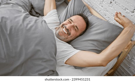 Beaming middle-aged man with grey hair joyfully stretching arms waking up in his cozy bedroom bed, relishing the comfort of a relaxing morning indoors - Powered by Shutterstock