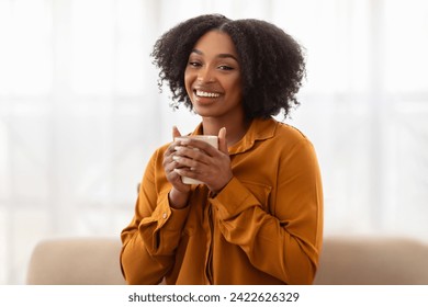 A beaming young african american woman with lush curly hair, dressed in a mustard-colored blouse, holds a mug while seated, exuding warmth and happiness in a softly lit room - Powered by Shutterstock