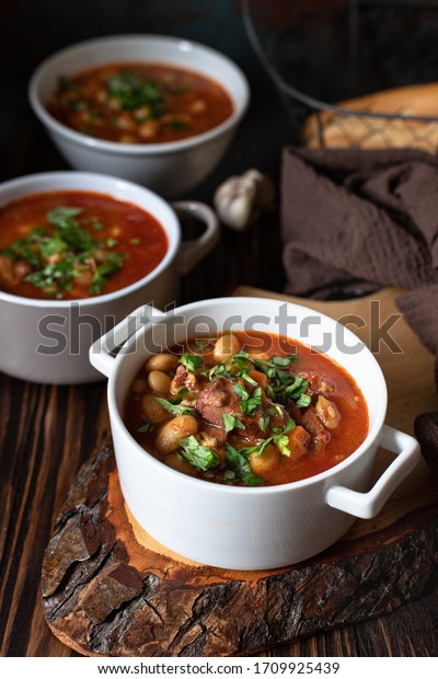 Bean soup with meat and\
vegetables served on a rustic board and wooden table with bread and\
garlic. Traditional Balkan soup Pasulj (Grah). Close-up, selective\
focus