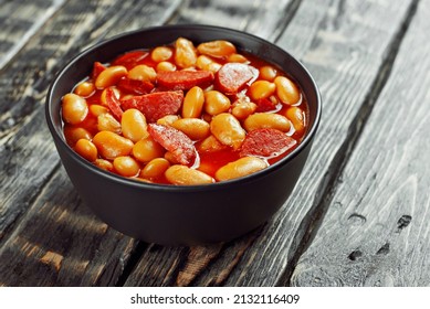 Beans baked with sausages. Beans with fried sausages in tomato sauce. Food in a black bowl on a brown wooden table. - Shutterstock ID 2132116409