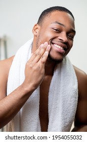 Beard care. Facial treatment. Male grooming. Happy satisfied shirtless African man with white towel enjoying touching dark face skin with hand after applying wax or oil.