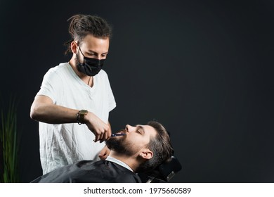 Beard Shaving In Barbershop. Barber With Dreadlocks In Black Medical Mask Trim Bearded Man At Quarantine Coronavirus Covid-19