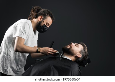 Beard Shaving In Barbershop. Barber With Dreadlocks In Black Medical Mask Trim Bearded Man At Quarantine Coronavirus Covid-19