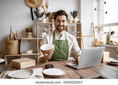 Bearded adult businessman engaged in retail trade of handmade tableware. Young Caucasian man potter happy with online sale of bowl on Internet against background of shelves with ceramics. - Powered by Shutterstock