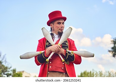 Bearded Adult Man In Red Suit Holding Bunch Of Clubs And Looking At Camera While Standing Against Cloudy Blue Sky During Show In Park