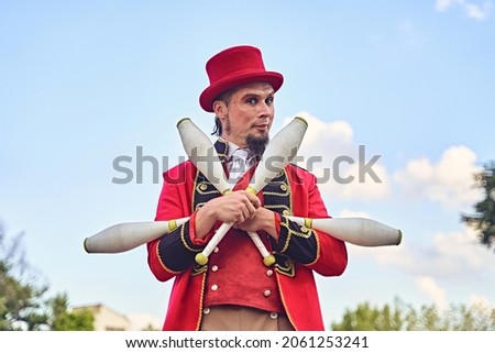 Bearded adult man in red suit holding bunch of clubs and looking at camera while standing against cloudy blue sky during show in park