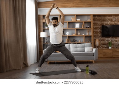 Bearded arabian man dressed in sport clothes standing on yoga mat with hands clenched above head during workout at home. Yoga practice for harmony, balance, soul and body care. - Shutterstock ID 2277805035