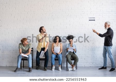 bearded art director calling brunette man near multiethnic actors sitting on chairs and waiting for casting