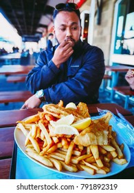 A Bearded Asian Guy Is Eating Fish And Chip Alfresco Near The Harbor.