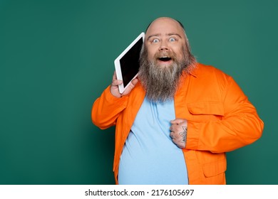 Bearded Astonished Man With Overweight Holding Digital Tablet Near Face Isolated On Green