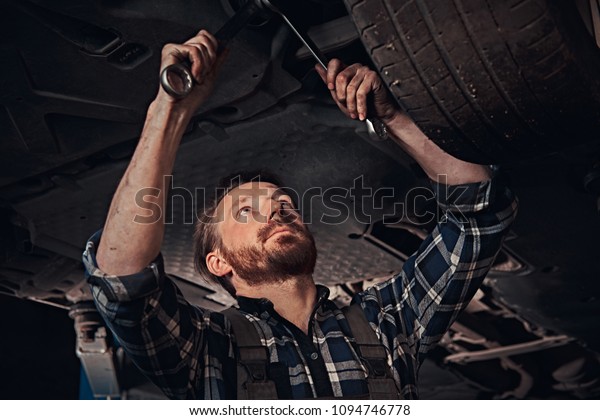 Bearded auto mechanic in a uniform repair the car\'s\
suspension with a wrench while standing under lifting car in repair\
garage. 