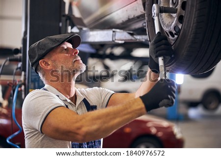 Bearded auto mechanic in work gloves using torque wrench while tightening bolts on wheel in automobile repair garage