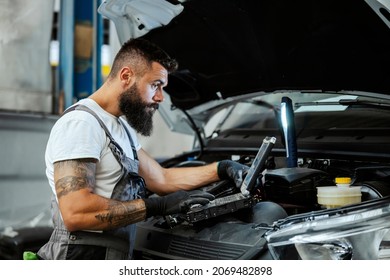 A bearded auto-mechanic in working uniform is standing next to a broken car and using the special laptop for car maintenance. An auto-mechanic in workshop repairs car - Powered by Shutterstock