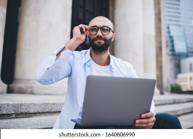 Bearded bald man in blue shirt working at laptop while relaxing on stairs adjusting glasses and looking up on blurred urban background