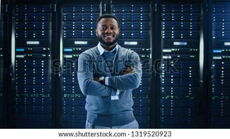 Bearded Black IT Engineer Standing and Posing with Crossed Arms in the Middle of a Working Data Center Server Room with Server Computers Working on a Rack.