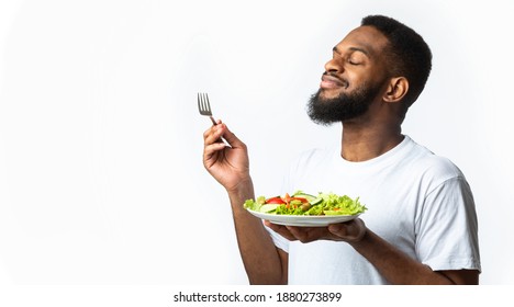 Bearded Black Guy Enjoying Delicious Vegetable Salad Standing With Plate Over White Studio Background. Healthy Nutrition, Dinner Delivery Concept. Side View, Panorama With Copy Space