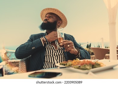 A bearded black man in a casual attire and a straw hat is adjusting his shirt lapel while drinking a glass of lemon iced tea accompanied by a vegetarian meal in an outdoor vegetarian restaurant - Shutterstock ID 2203905089