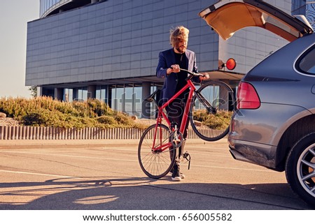 Bearded blond male with long hair holds fixed bicycle near the car with open trunk.