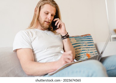 Bearded Blonde Man Talking On Cellphone While Working With Laptop At Home