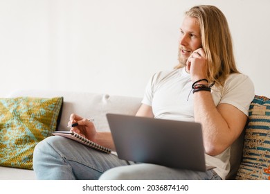 Bearded Blonde Man Talking On Cellphone While Working With Laptop At Home