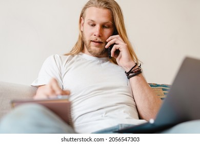 Bearded Blonde Man Talking On Cellphone While Working With Laptop At Home