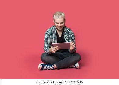 Bearded blonde man is using a tablet on the floor smiling on a red studio wall