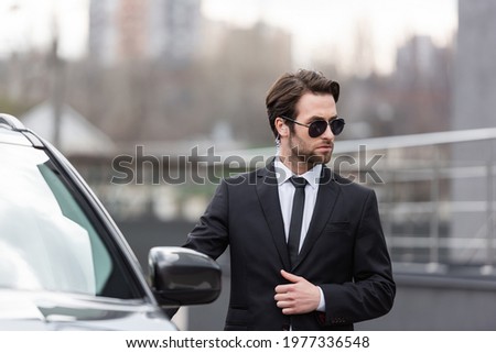bearded bodyguard in suit and sunglasses with security earpiece standing near modern car