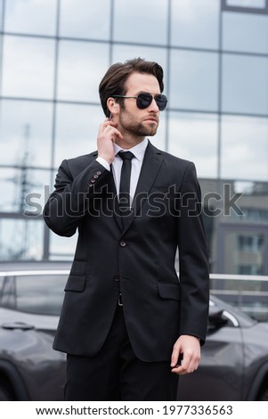 bearded bodyguard in suit and sunglasses with security earpiece near modern building on blurred background